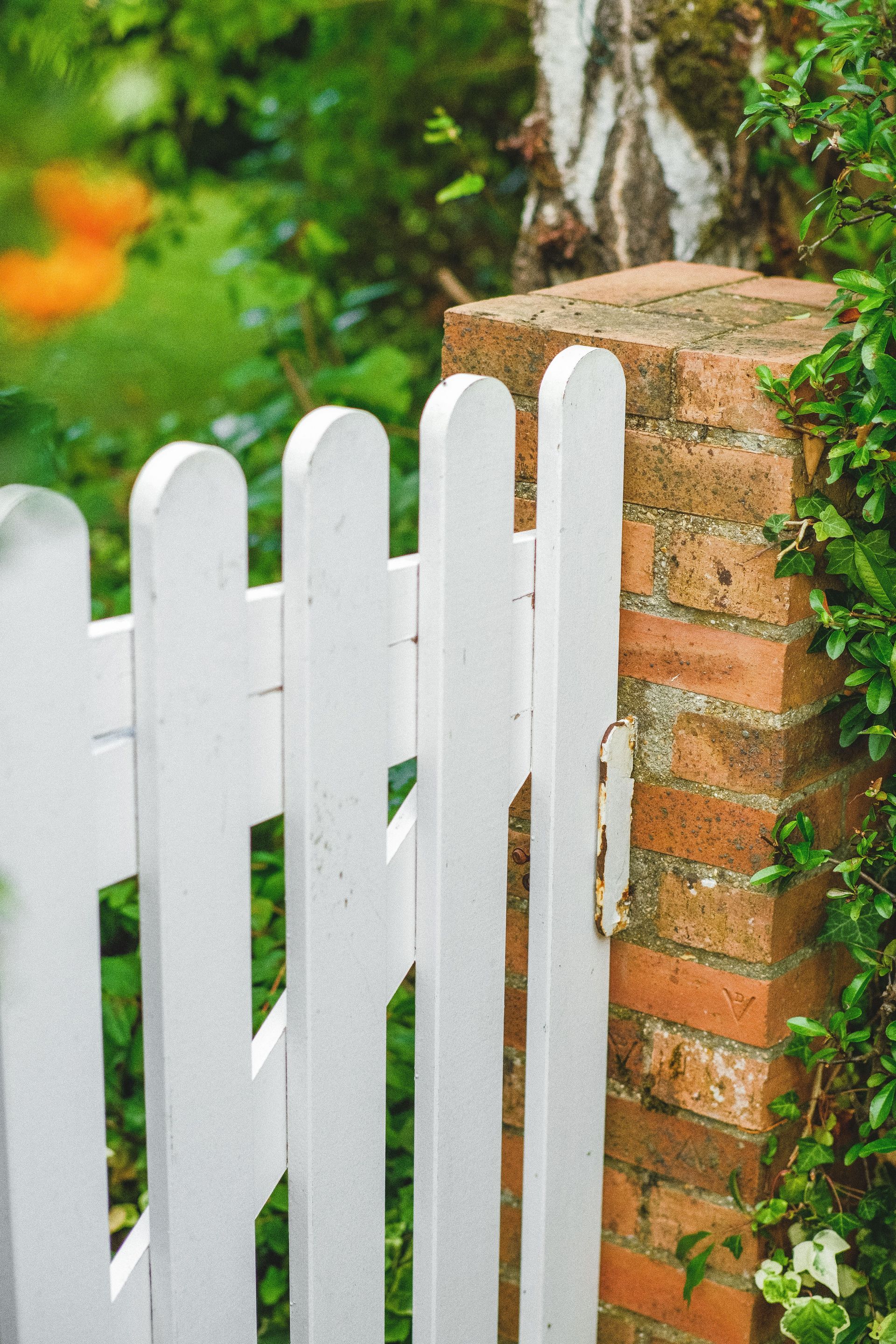 A serene garden entrance with a white wooden gate and red brick wall surrounded by lush greenery.